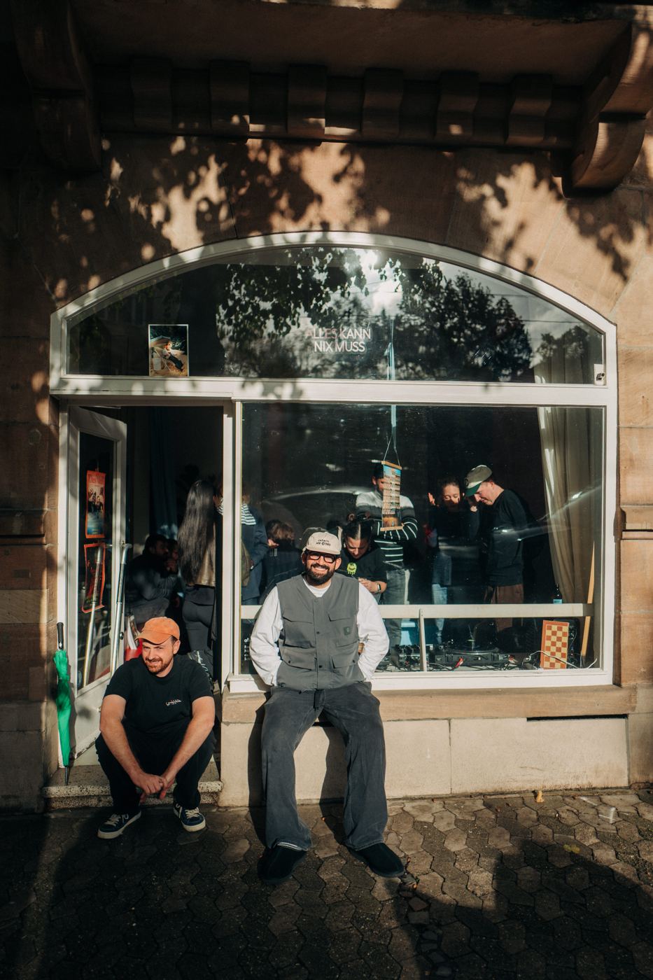 Zwei Männer sitzen lachend vor dem Schaufenster des Chess-Mates-Ladens, drinnen spielen Leute Schach