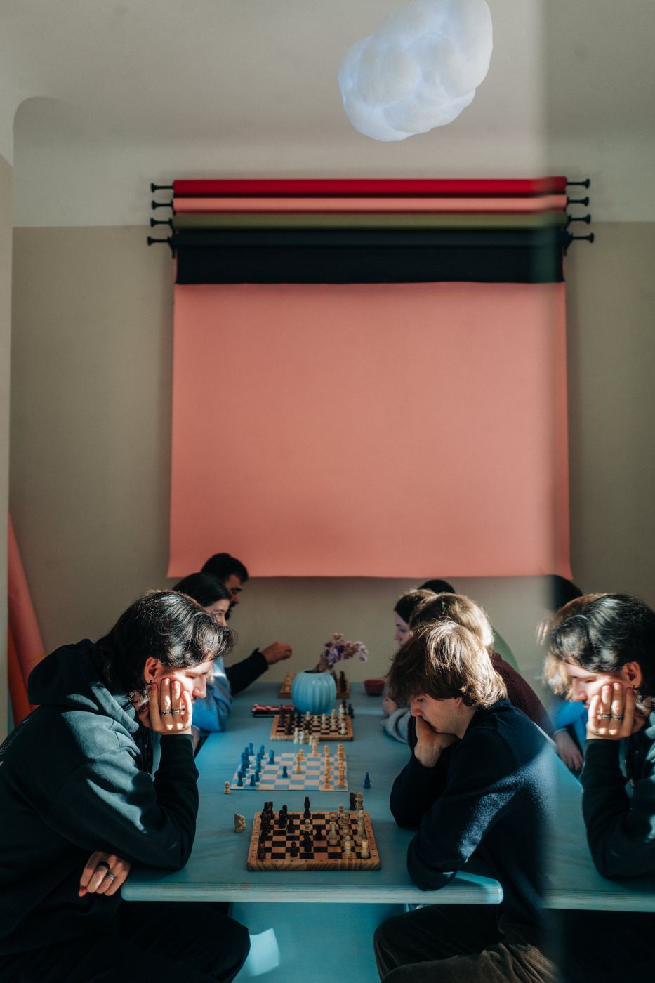Menschen spielen an einem langen hellblauen Tisch Schach vor einer rosa Wand
