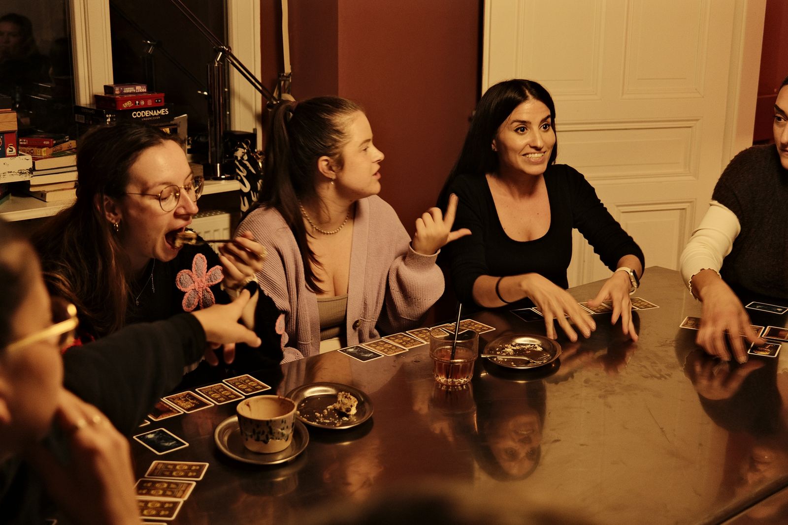 Gäste spielen Karten, eine Frau isst Kuchen, eine andere gestikuliert lachend